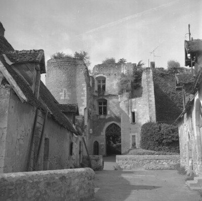 Czarno-białe zdjęcie pokazuje fragment brukowanej uliczki z małymi murowanymi domami po bokach. Na końcu uliczki brama wjazdowa do zamku ruin zamku.