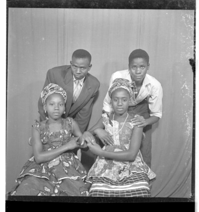 Two women wearing robes pagnes (wrapper dresses) and necklaces, sitting full frontal view in front of two men, one of whom is wearing a suit and the other is wearing a shirt and pants. Both are leaning over, facing forward. Taken in the studio.From Endangered Archives Programme EAP449, "Social history and cultural heritage of Mali"