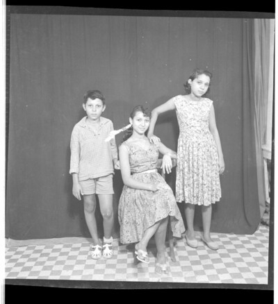 A woman wearing a dress sitting profile view between a boy wearing a shirt and shorts and a girl wearing a dress. The boy is standing full frontal view and the girl is standing profile view. Taken in the studio.From Endangered Archives Programme EAP449, "Social history and cultural heritage of Mali"