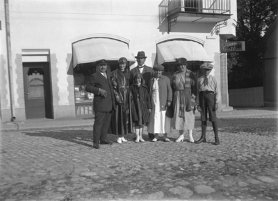 Kungsgatan i Nora.Grupp sju personer. I bakgrunden affärsbyggnader: Cigarrer och tidningar m.m., H .Hennberg Konditori.
