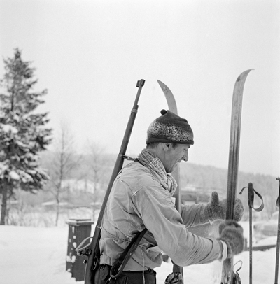 Fra det første offisielle kretsmesterskapet i skiskyting, som ble arrangert på Rasta ved Kongsvinger 8. januar 1961. Rennet gikk i bitende vinterkulde, det skal ha vært 29 kuldegrader da de 22 påmeldte løperne ble oppropt før start. Løypa var 8-9 kilometer lang og hadde fire standplasser, der det ble skutt på ulike hold (250, 150, 200 og 100 meter). På dette fotografiet ser vi Arvid Nyberg fra Trysil sette fra seg ski og staver. Han var kledd i en lys anorakk. På hodet hadde han ei rimete topplue og hendene var bare i kulda, rundt halsen et skjerf og på hendene et par digre ullvotter. Trysil-løperen fikk nest beste langrennstid i klasse A, men endte på sjetteplass fordi han bare traff 7 av 20 blinker i dette rennet. Nyberg var lagkamerat med toppløperen Hallgeir Brenden, og var med på å vinne tre stafettgull i langrenn for Vestre Trysil idrettsforening. Gjennom idretten kom han også inn i organisasjonslivet, og derfra kom han med i lokalpolitikken. Arbeiderpartimannen Arvid Nyberg var ordfører i Trysil fra 1972 til 1999, altså i 27 år. Han har seinere hatt et organisatorisk engasjement i Hedmark Jeger- og Fiskerforening.