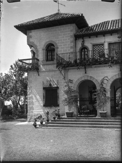 21 [Vista parcial desde el jardín del exterior de una casa de piedra con arcos  de medio punto en la entrada y piso superior abalconado. Macetas en las  escaleras de acceso] | Europeana